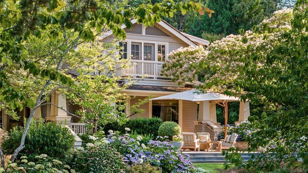 A leafy, landscaped view of a two-storey suburban house with a light weatherboard facade, gabled roof, upper-level balcony, covered ground-floor veranda, and abundant mature garden planting. The home has a warm, traditional residential character and is partially screened by trees and shrubs.