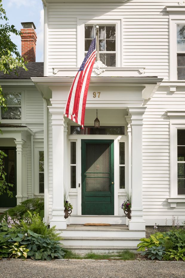 Front exterior view of a historic white clapboard home with an ornate covered entry porch, classical trim and columns, a dark green front door with sidelights, red brick chimney, and lush foundation planting. An American flag hangs above the entrance.