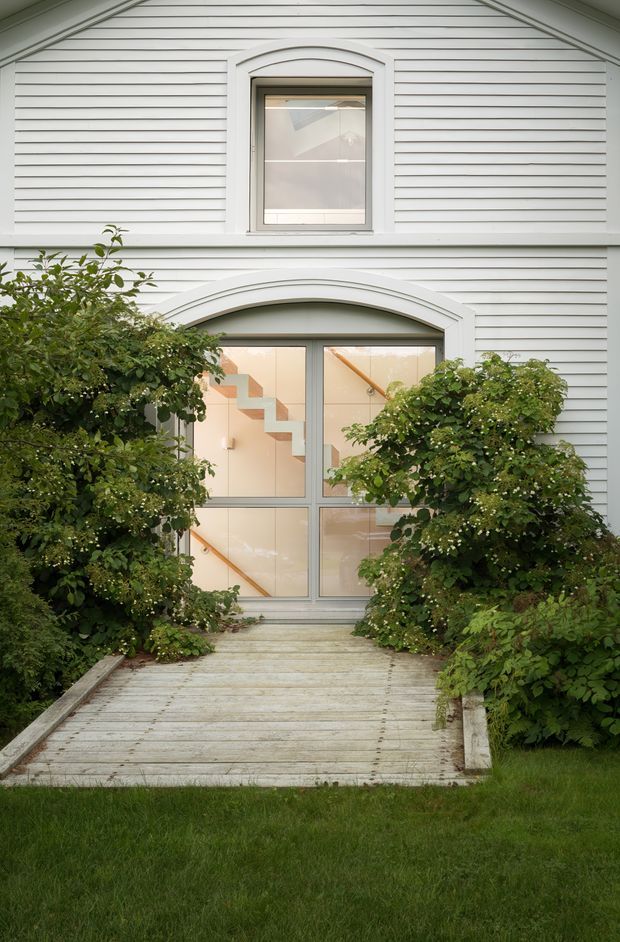 Exterior view of a light-painted, two-storey house with horizontal weatherboard cladding, decorative arched trim around the windows, and a timber walkway leading to large glazed doors. Dense shrubs frame the path, while a staircase is visible inside through the glazing.