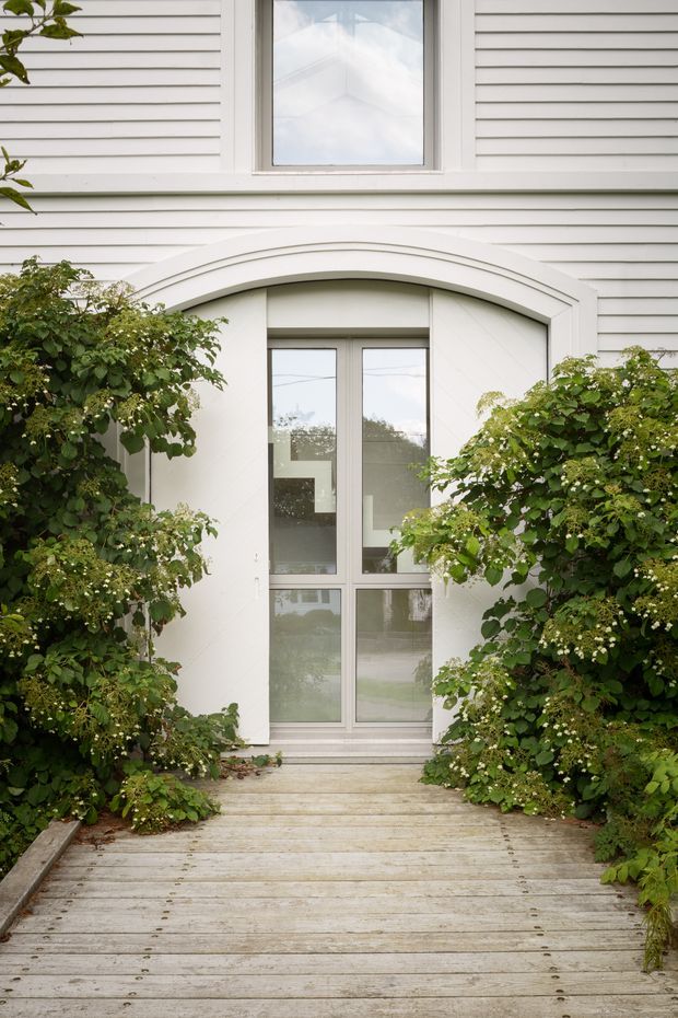 Exterior view of a white weatherboard-clad building entrance, framed by an arched architectural surround and glazed double doors. A timber boardwalk leads through dense green garden planting to the doorway, with a matching upper window above.