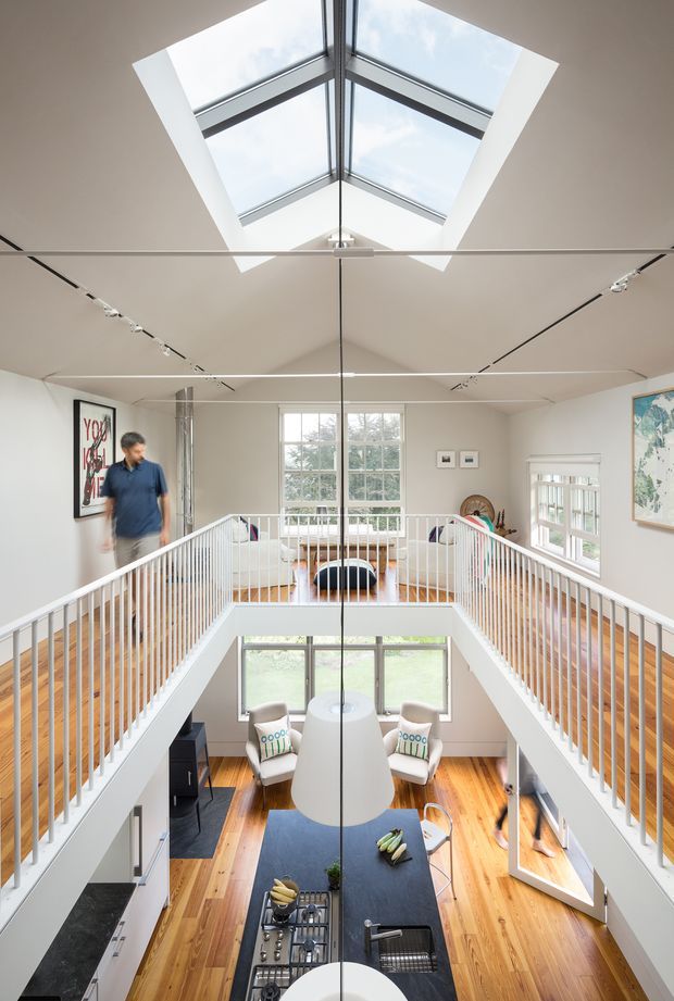 Bright contemporary multi-level interior centered on a double-height void with a mezzanine walkway and white balustrades. A large central skylight brings daylight into the space, while exposed timber flooring, white walls, extensive glazing, track lighting, and a sculptural pendant define the open-plan living area below. A seating area, dining or kitchen island, artwork, and upper lounge are visible.