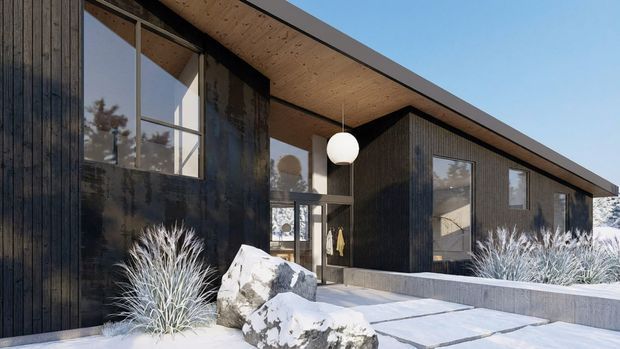 Exterior architectural view of a low, single-storey modern home in a snowy landscape. The building has dark vertical timber cladding, expansive black-framed glazing, a shallow mono-pitched roof with exposed timber soffit, and a covered entry with a spherical pendant light. Snow-covered planting, concrete paving, and large rocks frame the entrance.