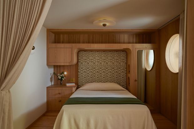 A compact, highly crafted bedroom with a single bed integrated into a warm timber built-in wall system. The bed has a textured woven headboard, layered cream and olive bedding, and is flanked by timber cabinetry and a small bedside drawer unit. Oval wall openings or mirrors appear on the right, while full-height drapery frames the left side. The room has a cozy, intimate atmosphere with warm ambient lighting and strong mid-century-inspired built-in detailing.