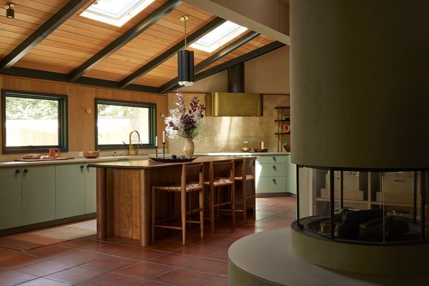 Warm, characterful kitchen with a central timber island and bar seating, muted green cabinetry, timber-framed windows, terracotta floor tiles, and a dramatic cylindrical fireplace in the foreground. The room has a timber-lined ceiling with exposed dark beams and multiple skylights, creating a strong indoor-outdoor connection and abundant natural light.