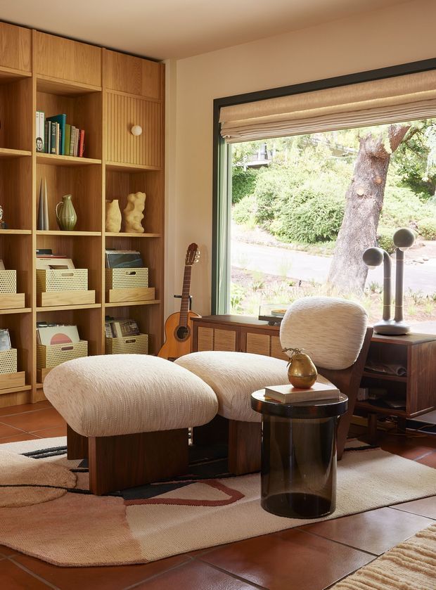 Warm, carefully styled living room or reading nook with a built-in timber library wall, integrated display shelving, and vertical timber panelling. Two sculptural upholstered lounge chairs with a matching ottoman face a small dark side table, while a guitar, decorative objects, books, and a pair of sculptural lamps add character. A large black-framed picture window with a woven Roman blind overlooks a leafy garden. Terracotta-toned floor tiles and layered organic-patterned rugs create a rich, earthy atmosphere.
