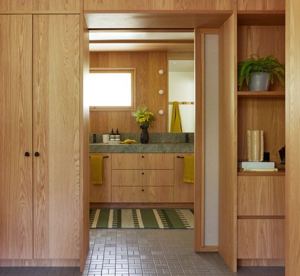 Warm, wood-lined bathroom viewed through a built-in timber storage and doorway composition. The room features a floating timber vanity with drawers, dark stone benchtop, wall-mounted round lights, a rectangular window, patterned tiled flooring, and a partially visible shower or bathing area. Integrated shelving on the right displays books and a potted plant, while a striped runner adds texture.