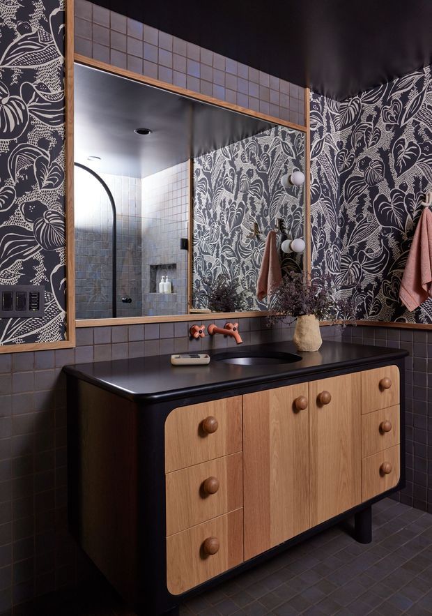 A moody, richly patterned bathroom vanity area. A large timber-framed mirror sits above a dark benchtop with a black basin and copper-toned wall-mounted tapware. The vanity has warm timber cabinetry with rounded wooden knobs, while dark blue-grey square tiles cover the walls and floor. Bold monochrome botanical wallpaper forms a feature wall, and towels and a small vase of dried foliage add texture.
