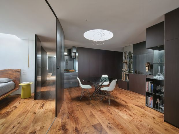 Open-plan contemporary interior combining a dining area, kitchen, living area and visible bedroom zone. Warm timber flooring contrasts with dark built-in cabinetry and a curved smoked-glass partition. The dining setting has a round glass table and modern white shell chairs beneath a sculptural circular pendant light. Dark joinery incorporates display shelving for glassware and objects, while the kitchen extends behind the partition. The space has a refined, minimalist contemporary character with warm wood and dark neutral finishes.