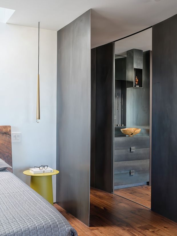 A contemporary bedroom interior featuring a partially visible bed, warm timber flooring, a small yellow side table, a slim pendant light, and extensive dark built-in wardrobe and cabinetry. Full-height mirrored wardrobe doors reflect additional dark joinery and drawers, while a sculptural timber bowl provides a small accent. The palette is restrained and moody, combining pale walls with charcoal-grey joinery and rich wood flooring.