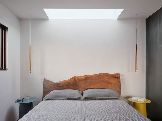 A contemporary bedroom viewed head-on, featuring a low-profile bed with a distinctive live-edge timber headboard, pale textured bedding, minimalist pendant lights, small round bedside tables, white walls, a dark feature wall, and a large rectangular skylight above the bed.