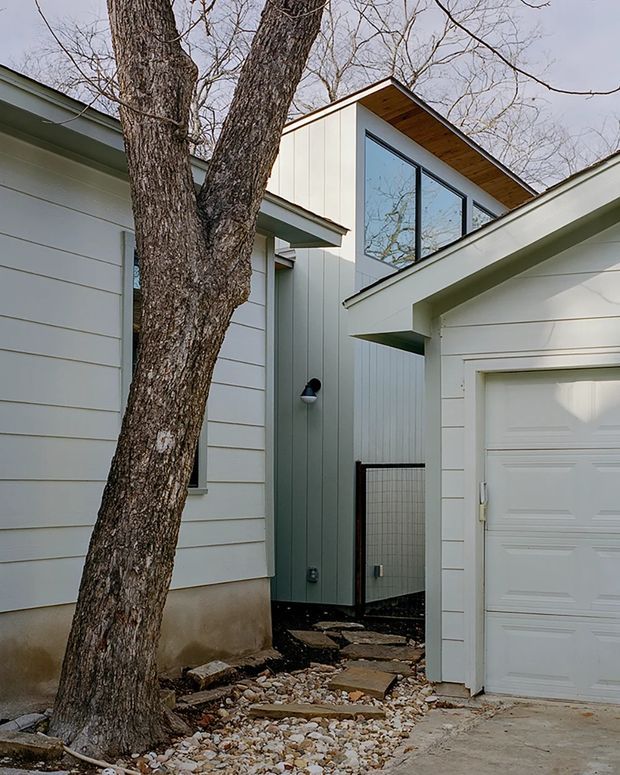Exterior view of a contemporary residential addition tucked among mature trees. The composition shows pale horizontal weatherboard cladding, a taller vertically clad volume with large black-framed glazing, a timber-lined roof soffit, and an attached white garage. A rocky landscaped walkway leads between the existing and new forms.