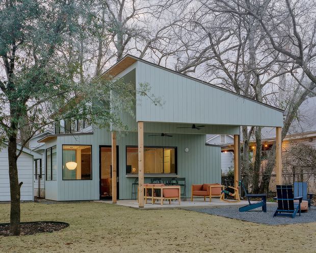 A compact single-storey modern pavilion-style house set among mature trees. The building has pale blue-grey vertical cladding, a dramatic sloping roof, large timber-framed windows and doors, and a covered outdoor verandah supported by timber posts. Outdoor seating and a small paved/gravel area sit beneath the canopy, with neighbouring residential structures partially visible.