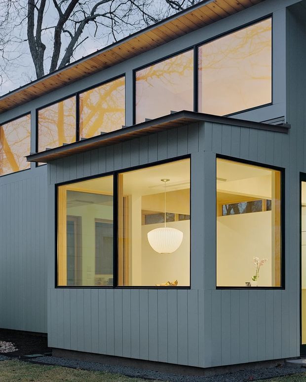 Exterior view of a contemporary two-storey house with a low-pitched roof, dark blue-grey vertical cladding, black-framed floor-to-ceiling windows, and warm timber soffits. Large glazed openings reveal a softly lit interior with a pendant light. Bare deciduous trees are reflected in the upper windows, and the building sits beside a gravel edge and lawn.