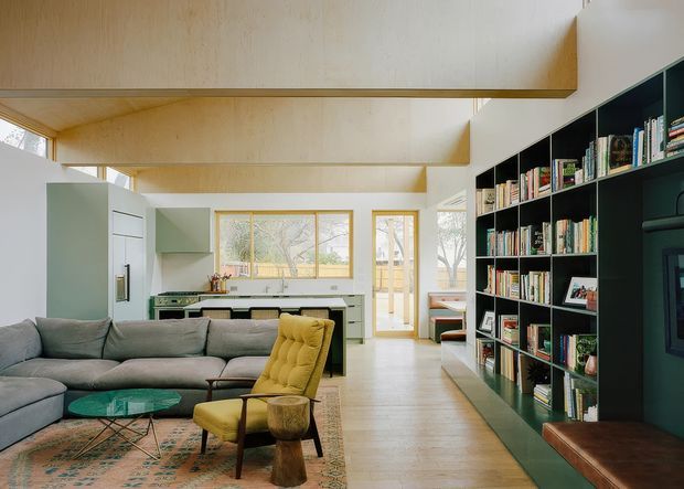 A bright, open-plan living area with a large grey sectional sofa, mustard lounge chair, patterned rug, glass coffee table, and extensive dark green built-in shelving filled with books. A compact kitchen is visible at the rear, with pale green cabinetry, timber-framed windows and doors, and abundant natural light. The space has a distinctive pale plywood-lined ceiling with deep exposed beams and a warm, contemporary mid-century-inspired character.