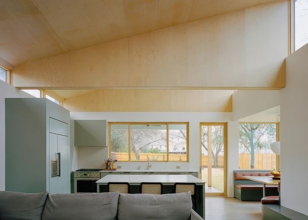 Open-plan contemporary kitchen and living area with a muted green kitchen, central island, integrated appliances, pale timber ceiling and upper wall surfaces, and extensive timber-framed glazing. A grey sofa occupies the foreground, while a built-in dining nook is visible to the right. The space has a warm, natural, architecturally detailed character with exposed timber surfaces and abundant daylight.