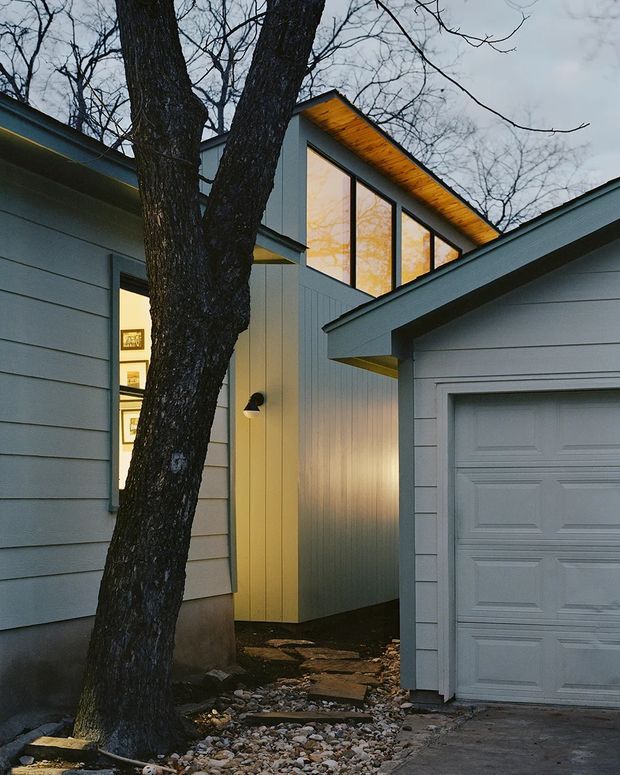 Exterior view of a compact contemporary residential composition among mature trees. Grey horizontal weatherboard-clad volumes surround a narrow illuminated entry passage, with a taller central volume featuring large vertical glazing and a warm timber-lined roof soffit. A detached or attached garage with a panelled garage door occupies the right side, while a stone-covered path and gravel landscaping lead toward the entry.