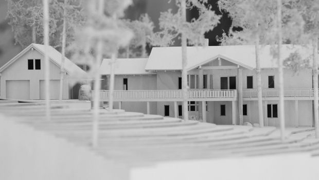 Monochrome architectural model or stylised exterior view of a low-rise residential complex with pitched gabled roofs, covered balconies, verandas, repeated windows, and attached garage-like structures. Trees and stepped foreground elements surround the building.