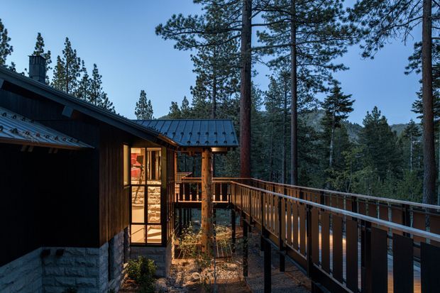 Exterior view of a dark timber-clad mountain cabin with a low pitched metal roof, stone base, warm-lit glazing, and an elevated timber deck and walkway extending through a pine forest.