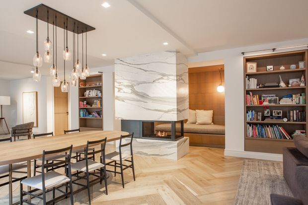 Open-plan contemporary living and dining area with a long light timber dining table, black-framed chairs, a dramatic cluster pendant light, built-in bookshelves, and a central marble-clad fireplace dividing the dining space from a warm built-in seating nook. The room has pale herringbone timber flooring, white walls and ceiling, recessed lighting, and dark timber shelving with a refined, warm-neutral palette.