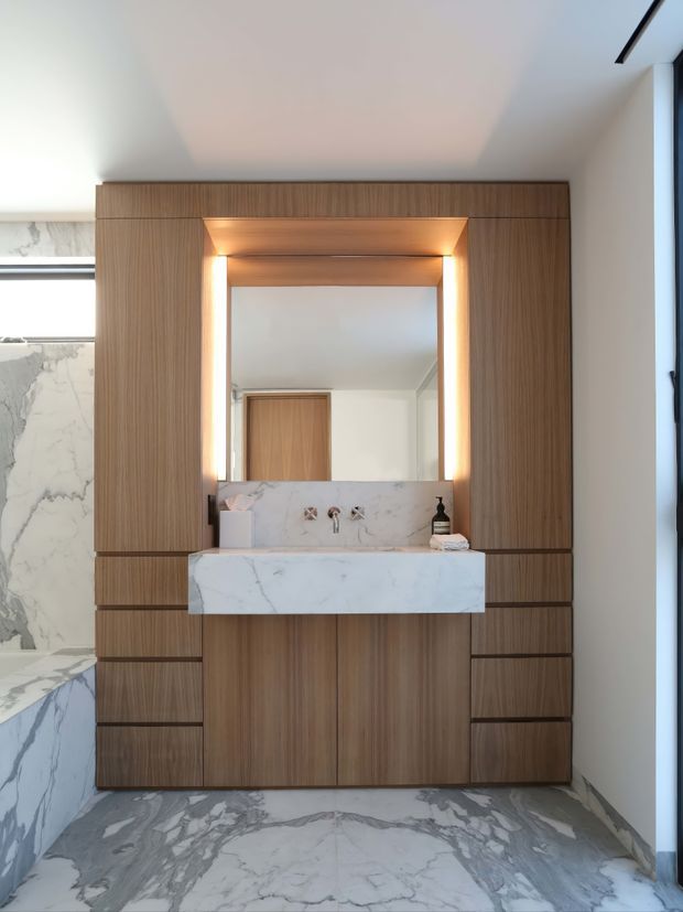 Contemporary bathroom vanity wall featuring extensive warm wood cabinetry, a white marble benchtop and integrated splashback, a large illuminated mirror, and large-format marble wall and floor tiles. The floating vanity has symmetrical drawer storage and a sculptural stone basin area, creating a refined, luxurious appearance.