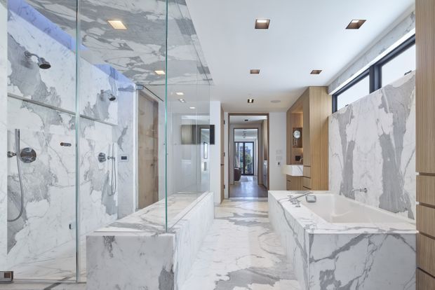 A bright, luxurious contemporary bathroom with extensive white marble-look wall and floor surfaces, a freestanding bathtub in the foreground, a glass-enclosed shower area on the left, and a long timber vanity with integrated storage on the right. Large windows provide natural light, while recessed ceiling lights and a corridor beyond extend the view through the home.