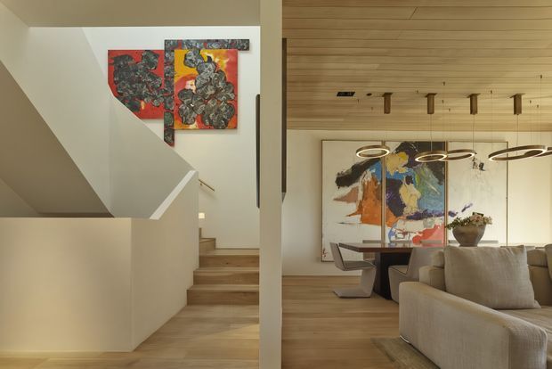 Warm contemporary interior showing a timber-lined open-plan living and dining area beside a stairway. The stair has painted light walls and timber treads, with colourful abstract artwork on the wall. The living space contains a large neutral sectional sofa, dining table and sculptural chairs, pendant lighting, and a large abstract artwork. Vertical timber wall and ceiling lining, recessed lighting, and restrained furnishings create a refined, natural atmosphere.