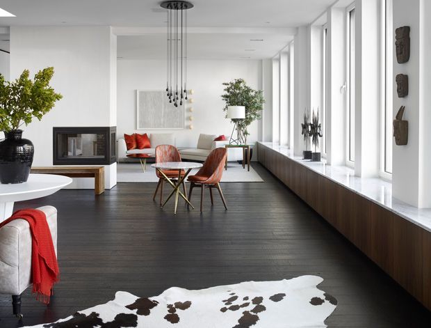 A bright, spacious contemporary living room with a long wall of floor-to-ceiling windows, dark timber flooring, and a built-in dark wood window seat or storage unit. The room includes a fireplace, sofas and sculptural lounge chairs, a small round coffee table, a tall linear pendant light, indoor plants, and restrained decorative wall art. The palette combines white walls, dark timber, neutral upholstery, and red accent textiles.