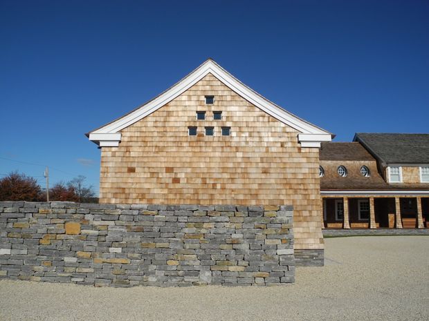 Exterior view of a shingle-clad gable-front building behind a low dry-stacked stone wall. The facade features white-trimmed roof edges and small square windows arranged decoratively near the gable peak, with a connected darker shingle-roofed wing and covered entry visible to the right. A gravel forecourt and clear blue sky surround the structure.