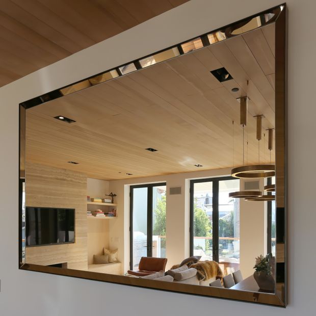 A large decorative wall mirror reflects a warm contemporary living room with a timber-lined ceiling, built-in timber television cabinetry, shelving, lounge seating, pendant lights, and generous glazed doors and windows opening toward an outdoor area. The mirror has a dark metallic frame with faceted detailing along the top and sides.
