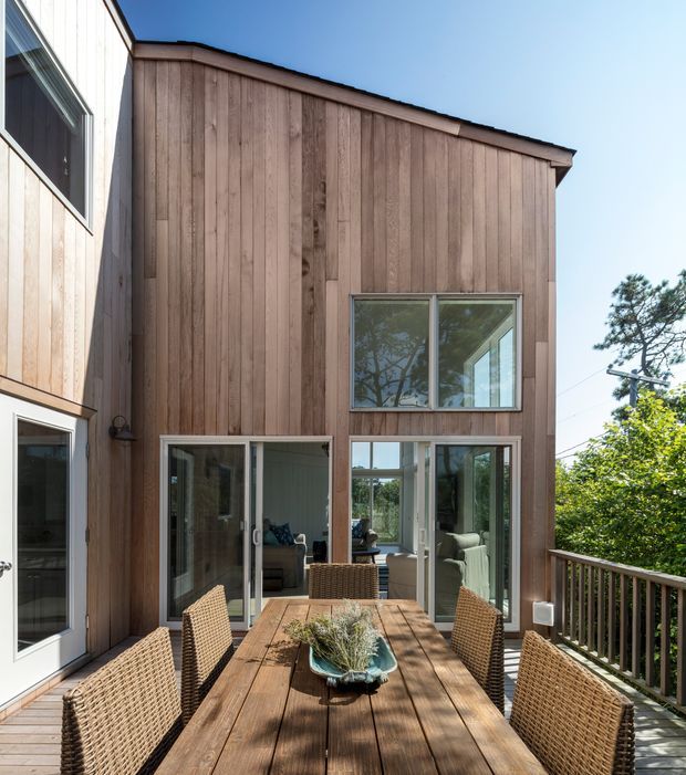 Exterior view of a modern timber-clad house centered on a raised outdoor dining deck. The facade combines vertical natural wood cladding with large glazed doors and windows, while a timber dining table, woven chairs, and deck balustrade occupy the foreground. The image is sunlit with views of trees beyond the terrace.