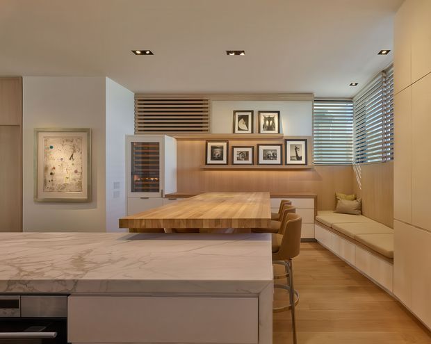 A warm, contemporary open-plan kitchen and dining space with a large pale marble island in the foreground, timber cabinetry and flooring, a substantial timber dining table, and integrated upholstered bench seating along the window wall. Horizontal timber screens and venetian blinds filter light, while recessed ceiling spotlights and framed artwork add understated detail.