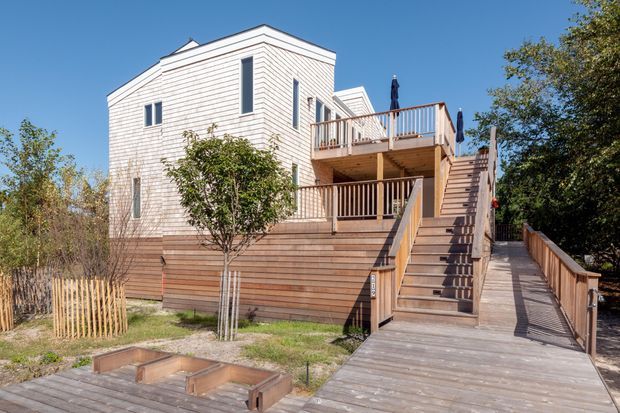 Exterior view of a multi-level timber-clad house with a raised deck, broad outdoor staircase, timber boardwalk, and upper balcony. The light shingle-style wall cladding contrasts with warm horizontal timber decking and retaining walls, set among established landscaping under a clear blue sky.