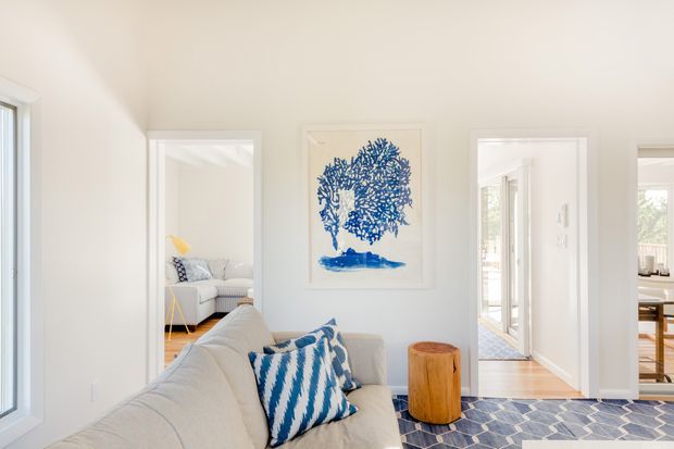 Bright, airy living room with a cream upholstered sofa, blue-and-white patterned cushions, a geometric blue rug, and a small timber side table. White walls, ceiling, trim, and doorways create a clean neutral backdrop, with views into adjoining rooms and a blue botanical artwork as the main decorative feature.