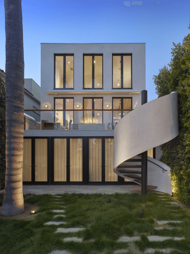 Twilight exterior view of a narrow, multi-storey contemporary residence with a clean white rendered facade, tall black-framed windows and doors, warm interior lighting, a glazed upper terrace, and a prominent sculptural white spiral staircase projecting into the landscaped front garden. A palm tree, hedging, stepping stones, and subtle garden lighting frame the building.