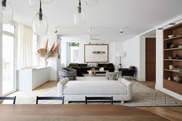 Bright, spacious open-plan living room viewed from a dining area, featuring white walls and ceiling, pale timber flooring, a large textured area rug, a white upholstered sofa, dark lounge seating, a central coffee table, extensive glazing with sheer curtains, pendant lights, and custom dark timber display shelving and cabinetry. The room has a refined contemporary feel with neutral tones and natural textures.