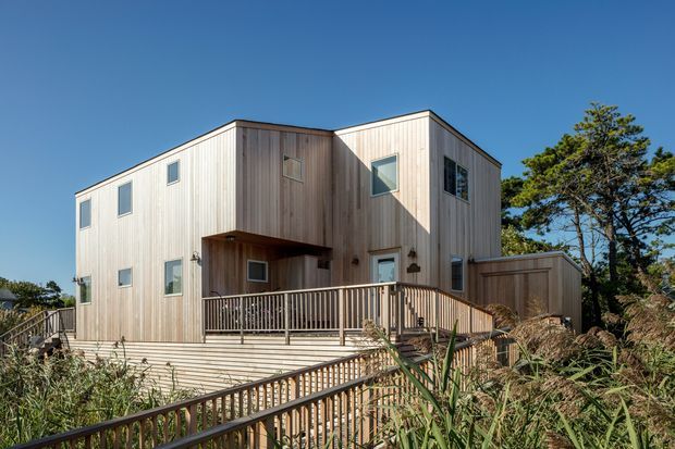 Exterior view of a modern multi-level house clad in light vertical timber weatherboards, with simple rectangular volumes, small square windows, projecting entry canopy, and extensive timber decking and railings. The home is set among coastal-style vegetation beneath a clear blue sky.