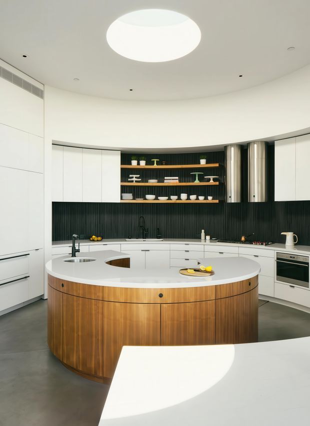 A high-end contemporary kitchen with a large curved central island featuring vertically grained timber cabinetry and a white stone benchtop. The surrounding kitchen has extensive white cabinetry, integrated appliances, a dark vertically ribbed feature wall, open timber shelving with crockery and decor, and a sculptural circular ceiling light. The space has a clean, refined, minimalist appearance with strong curved forms and a polished architectural fit-out.