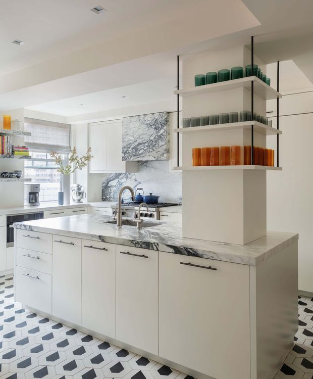 Bright contemporary kitchen centered on a large island with white cabinetry, dark linear hardware, and a dramatic veined marble benchtop and splashback. A suspended open shelving unit displays coloured glassware, while geometric black, white, and pale-blue patterned floor tiles add visual interest. The room includes integrated appliances, a sink and tapware, pendant-free recessed lighting, and generous window glazing.