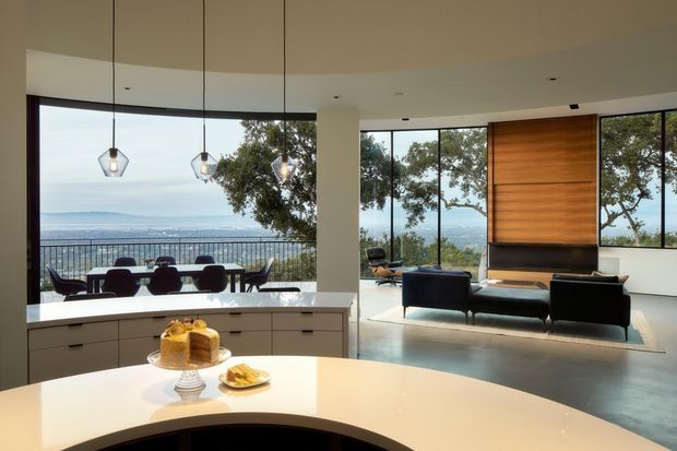 Open-plan contemporary kitchen and living area with a large curved light benchtop in the foreground, streamlined pale cabinetry, pendant lights, and a dining area beside full-height glazing. The adjoining living room has dark sofas and armchairs arranged around a fireplace set into a warm timber feature wall. Expansive windows open to a distant coastal or urban view and bring in abundant natural light.