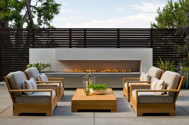 A refined outdoor lounge terrace with two pairs of cushioned wooden armchairs arranged around a low timber coffee table. The space is enclosed by a dark horizontal slatted privacy screen and features a long linear gas fireplace set into a pale rendered surround, with landscaped greenery visible around the edges.