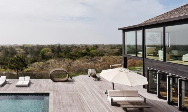 Exterior view of a contemporary multi-level home with dark timber cladding, extensive floor-to-ceiling glazing, and a broad timber deck surrounding a swimming pool. Outdoor loungers, a sculptural chair, and a large umbrella create a relaxed terrace setting, while the elevated house overlooks a wooded landscape.