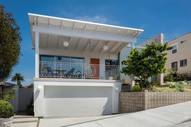 A bright contemporary two-storey residential facade viewed from the driveway. The home features a white rendered exterior, a projecting flat roof with exposed white rafters, a covered upper-level balcony with slender metal balustrades, large glazed doors, and an integrated double garage. Concrete paving, a tiled retaining wall, and landscaped planting frame the frontage.