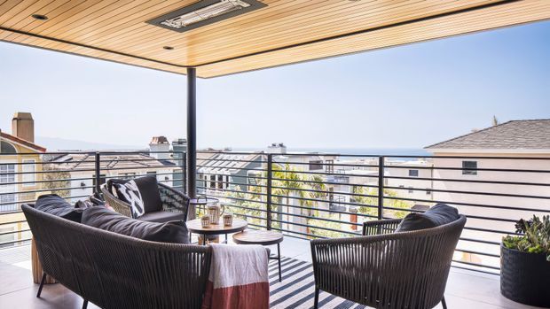A covered outdoor terrace or balcony furnished with dark woven lounge chairs and a small round coffee table. The space has a warm timber slatted ceiling with recessed lighting, slim dark structural posts, horizontal metal balustrades, and views across neighbouring rooftops toward a bright distant horizon. Potted planting adds greenery to the relaxed entertaining area.