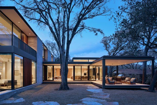 A dusk exterior view of a large contemporary residence arranged around a landscaped courtyard. The home has strong horizontal and rectilinear forms, extensive floor-to-ceiling glazing, dark metal-framed windows and doors, warm timber-clad wall sections, and a covered outdoor living pavilion with lounge furniture. A mature deciduous tree and irregular stone pathway occupy the foreground.