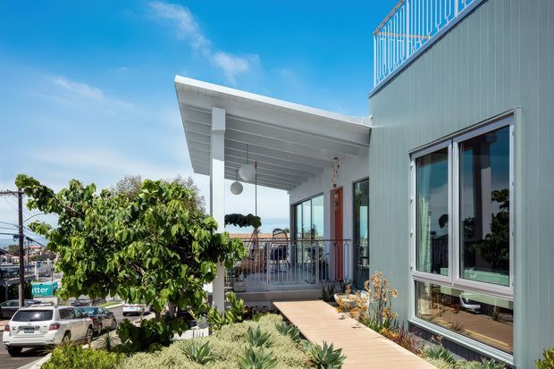 Exterior view of a contemporary multi-level residence with pale grey vertical cladding, large glazed windows, a projecting covered entry porch, metal balcony railings, and a timber walkway through drought-tolerant landscaping. The elevated setting provides glimpses of neighbouring buildings and a blue sky beyond.