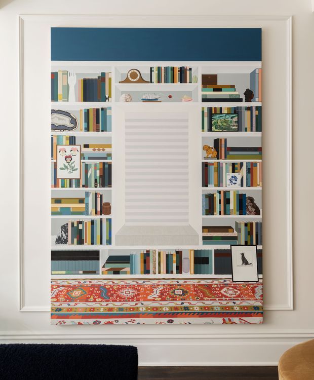 A decorative built-in-style bookshelf wall with white shelving, a large recessed central panel, and a deep blue upper section. The shelves are filled with colourful books, framed artwork, small sculptures, and objects. A vivid patterned textile spans the lower portion, creating a layered, eclectic focal point. White traditional trim surrounds the feature, with glimpses of dark timber flooring and upholstered furniture at the bottom edge.