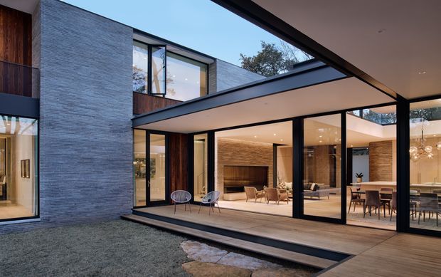 Exterior architectural view of a contemporary two-storey residence organized around a landscaped courtyard. The home features expansive black-framed glazing, warm timber wall panels and decking, pale textured masonry cladding, deep cantilevered roof planes, and a partially glazed roof or clerestory opening. Interior living and dining areas are visible through the glazing.