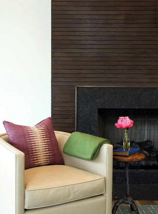 Close-up interior view of a refined living room seating area. A cream upholstered armchair with a burgundy patterned cushion and green throw sits beside a dark fireplace framed by horizontally laid dark timber wall panelling. A small round side table holds books and a pink flower arrangement. The composition is warm, richly textured, and intimate.