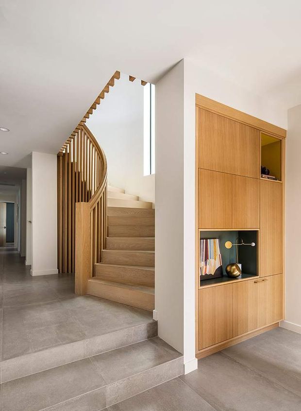 Bright contemporary residential entry hall featuring a sculptural timber staircase with curved vertical slat balustrade, pale timber stair treads, large-format grey floor tiles, white walls, and a full-height timber built-in display cabinet with recessed niches. A narrow vertical window brings natural light into the stairwell.