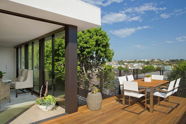 A contemporary flat-roofed home with a timber outdoor dining deck overlooking a suburban landscape. The deck has a timber-clad floor, dark horizontal privacy balustrade, and a dining table with white chairs. Full-height glazed sliding doors open to a bright interior sitting area, framed in dark metal. Lush trees and distant houses are visible beyond the terrace.